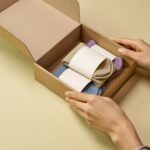 Solutions A woman's hands packing colorful socks into a cardboard box on a light yellow surface.